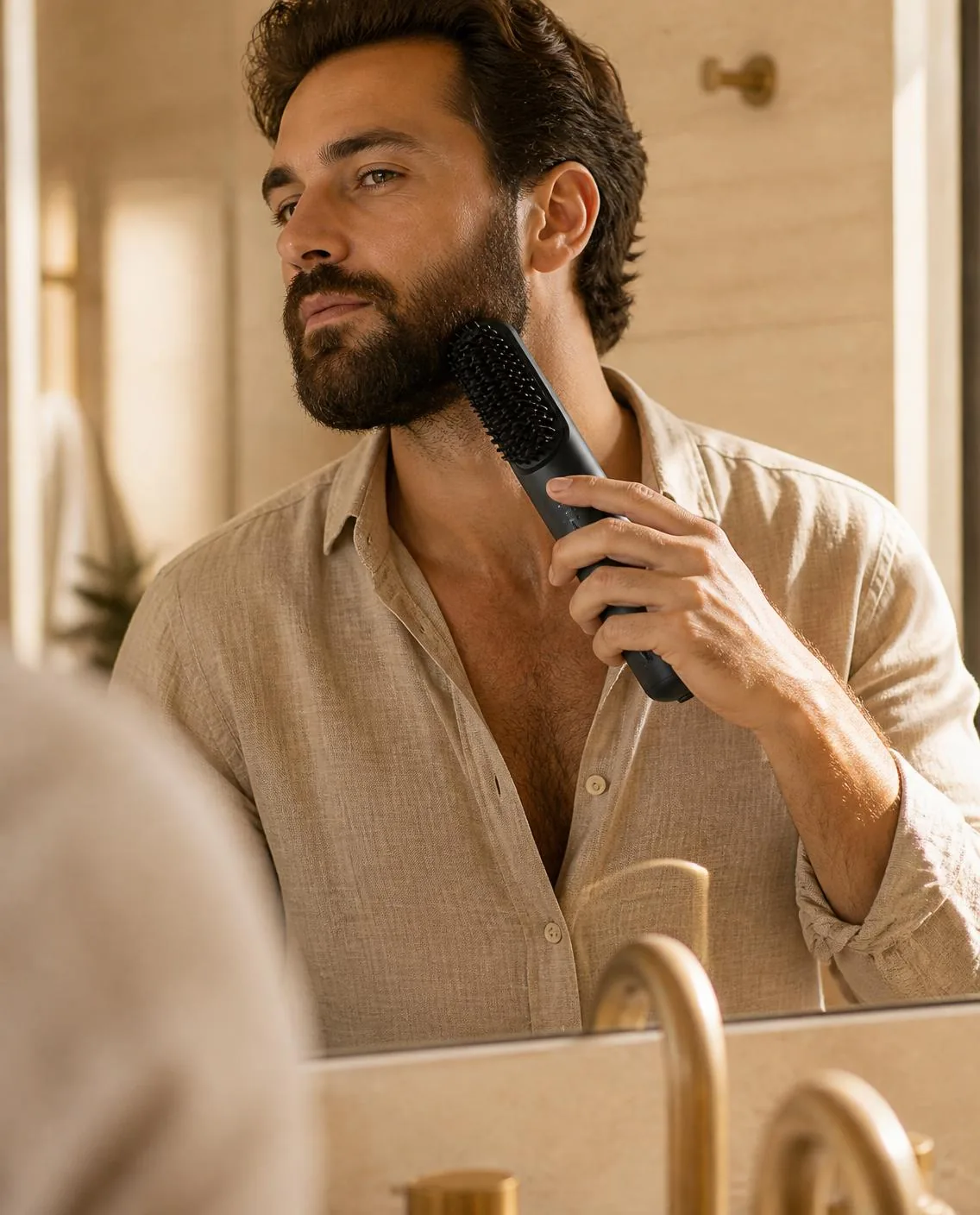 A bearded man in a linen shirt running the black cordless brush through his beard at the mirror